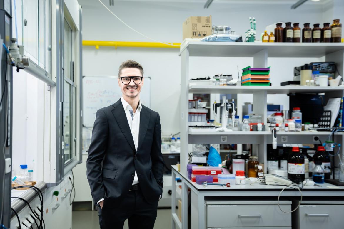 Zinātnieks uzvalkā stāv savā laboratorijā starp materiāliem