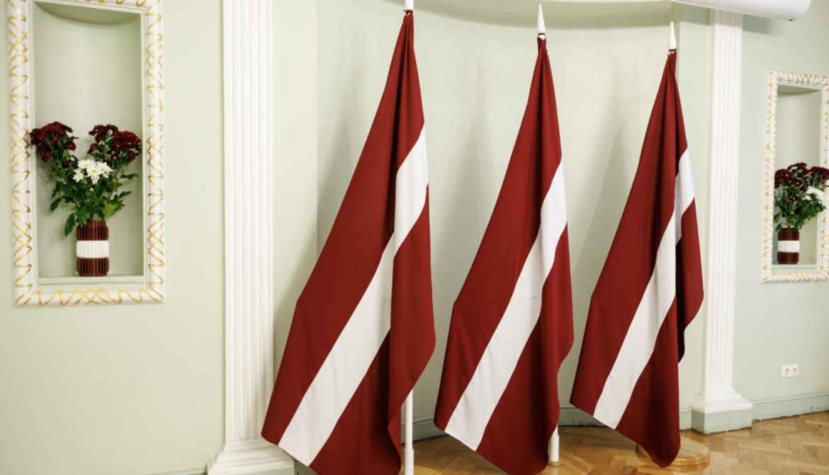 Trīs lieli Latvijas karogi, uz plaukta ziedi Latvijas karoga krāsās